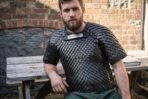 Blackened steel scale armour shirt with a rune plaque at the throat worn by a bearded man seated at a garden table