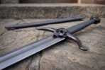 Close up of a blackened steel skull sword guard with horned skull and chevron etched blade on stone paving, black scabbard behind