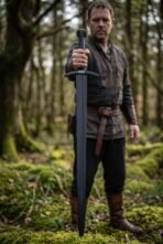 Blackened steel sword held point down by a bearded man in a mossy forest, the curved guard at his chest