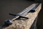 Blackened steel sword with fullered blade, black leather grip and disc pommel resting on an old wooden fence rail