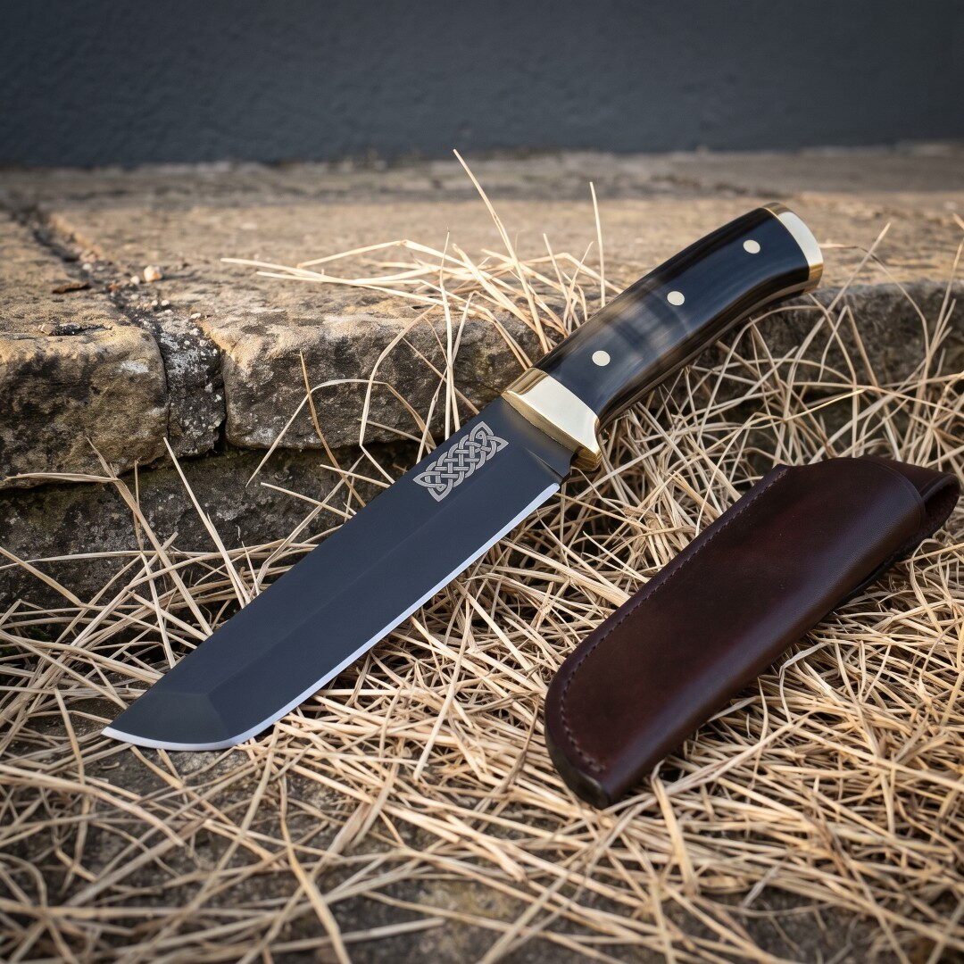 Blackened tanto knife with an etched Celtic knot panel, black buffalo horn scales and brass bolster lying on dry straw b Blackened tanto knife with an etched Celtic knot panel, black buffalo horn scales and brass bolster lying on dry straw beside a dark brown leather belt sheath