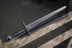 Blackened Viking sword with ball pommel and black corded grip laid along a weathered fence rail next to its black scabbard