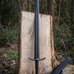Blackened yggdrasil sword standing point down against a cut log in woodland showing its tree of life pommel, cord wrapped grip and black leather scabbard