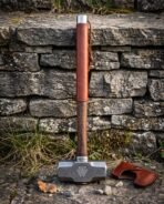 Blacksmith hammer with a Damascus patterned double faced head cut with a valknut, hardwood handle wrapped in leather, standing against a dry stone wall.