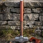 Blacksmith hammer with a Damascus patterned double faced head cut with a valknut, hardwood handle wrapped in leather, standing against a dry stone wall.