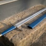 Blue leather Viking sword in 1095 steel with a faceted brass pommel and knotwork etched blade lying on a stone step beside its matching blue leather scabbard