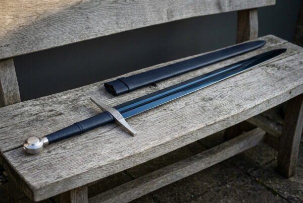Blued arming sword with a bright polished wheel pommel and a knotwork etched guard, resting on a weathered garden bench beside its black leather scabbard
