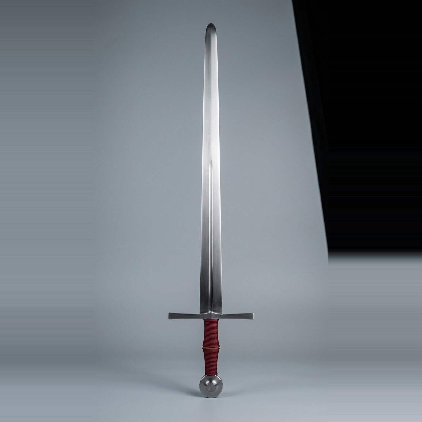 Haldvane blunt arming sword trainer Blunt arming sword trainer with a broad fullered blade, straight cross and burgundy cord grip with a tan riser standing point up against a plain grey studio backdrop