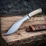 Bone handle kukri knife in satin polished high carbon steel with rune etched spine, three pin scales, steel bolster and brown leather sheath resting on a split log