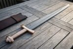 Bone handle Viking sword in pattern welded Damascus steel with rune etched fuller and brass lobed pommel laid beside a brown leather scabbard on decking