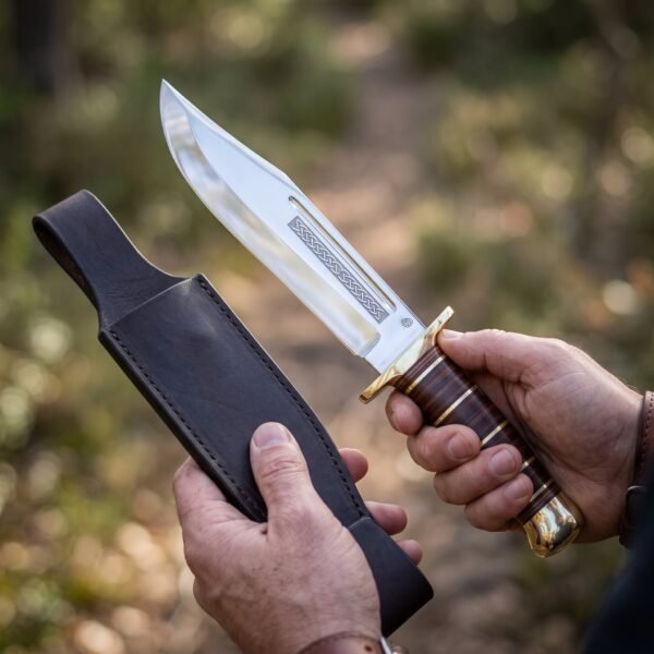Bowie knife with stacked leather handle, brass double guard and knotwork etched clip point blade beside a dark leather sheath