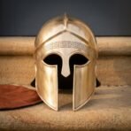Brass Greek hoplite helmet with crest ridge, nasal and long cheek pieces shown face on beside a leather pad on stone