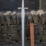 Brass hilt arming sword with a wheel pommel and pale polished grip standing point down against a dry stone wall beside its tan leather scabbard