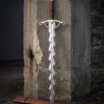 Brass hilt flamberge sword with a wavy flame blade in satin 1095 steel, ring terminal guard and faceted brass pommel standing against an old stone doorway
