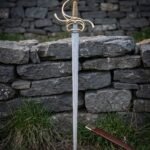 Brass hilt rapier with a folded Damascus blade standing point down in grass before a dry stone wall next to its brown leather scabbard