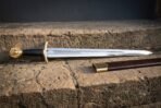 Brass hilted lion head short sword with a knotwork etched fuller and brown leather scabbard laid on warm sandstone blocks in low evening light