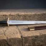 Brass hilted lion head short sword with a knotwork etched fuller and brown leather scabbard laid on warm sandstone blocks in low evening light