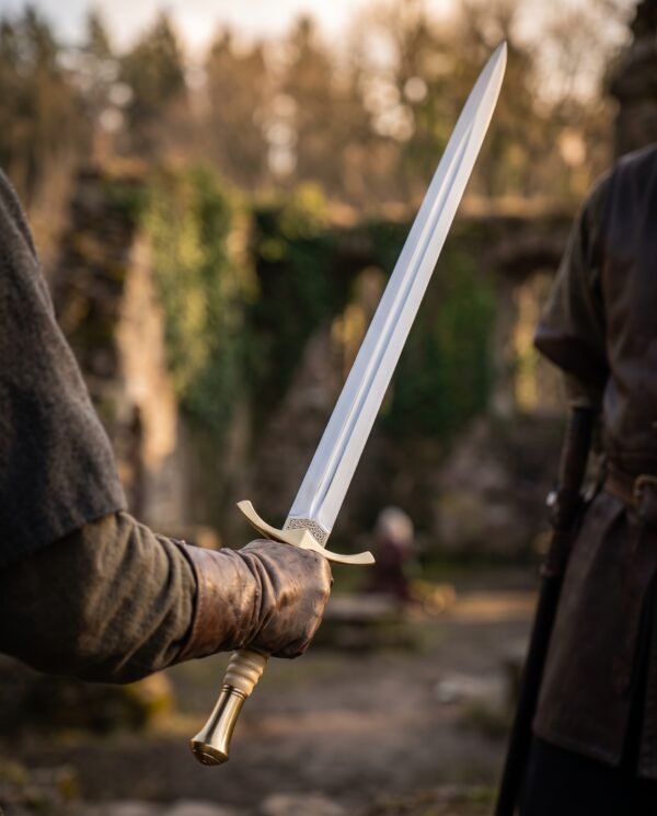 Brass hilted steel sword held in a gloved hand, fullered blade raised beside ivy covered stone ruins in evening light