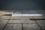 Bright polished 1095 steel wolf head longsword with a long black leather grip and etched knotwork at the ricasso lying on grey paving slabs beside a brown scabbard