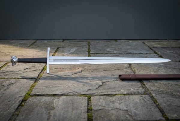 Bright polished 1095 steel wolf head longsword with a long black leather grip and etched knotwork at the ricasso lying on grey paving slabs beside a brown scabbard