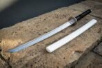 Bright polished white saya wakizashi in clay quenched 1095 steel showing a wave hamon along the edge, laid on a sunlit sandstone ledge with its gloss scabbard