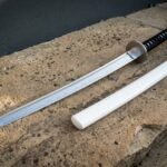 Bright polished white saya wakizashi in clay quenched 1095 steel showing a wave hamon along the edge, laid on a sunlit sandstone ledge with its gloss scabbard