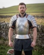 Bright steel cuirass with articulated pauldrons, hooped fauld and rune plaque worn by a man in a dark tunic in front of a dry stone wall