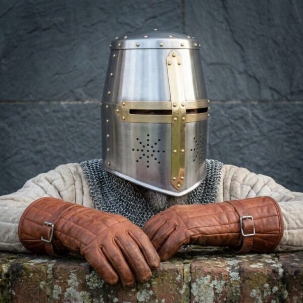 Bright steel great helm with brass cross worn by a man in a chainmail coif and quilted tunic, brown leather gloves folded in front