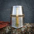 Front view of a bright steel great helm with brass cross reinforce, etched knotwork sights and drilled breaths on a lichen covered brick wall