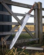 Broad bladed 1095 steel arming sword leaning across a weathered timber field gate in low evening light with its cream scabbard lying in the grass