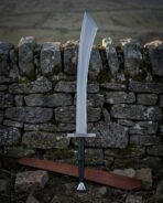 Broad curved viking scimitar with a knotwork engraved crossguard and black leather grip standing against a dry stone wall above a long brown leather scabbard