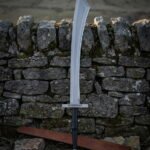 Broad curved viking scimitar with a knotwork engraved crossguard and black leather grip standing against a dry stone wall above a long brown leather scabbard
