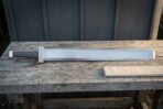 Broad hand forged viking sword with a Celtic knotwork guard and block pommel lying on a weathered wooden bench beside a linen wrapped scabbard
