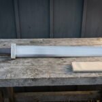 Broad hand forged viking sword with a Celtic knotwork guard and block pommel lying on a weathered wooden bench beside a linen wrapped scabbard