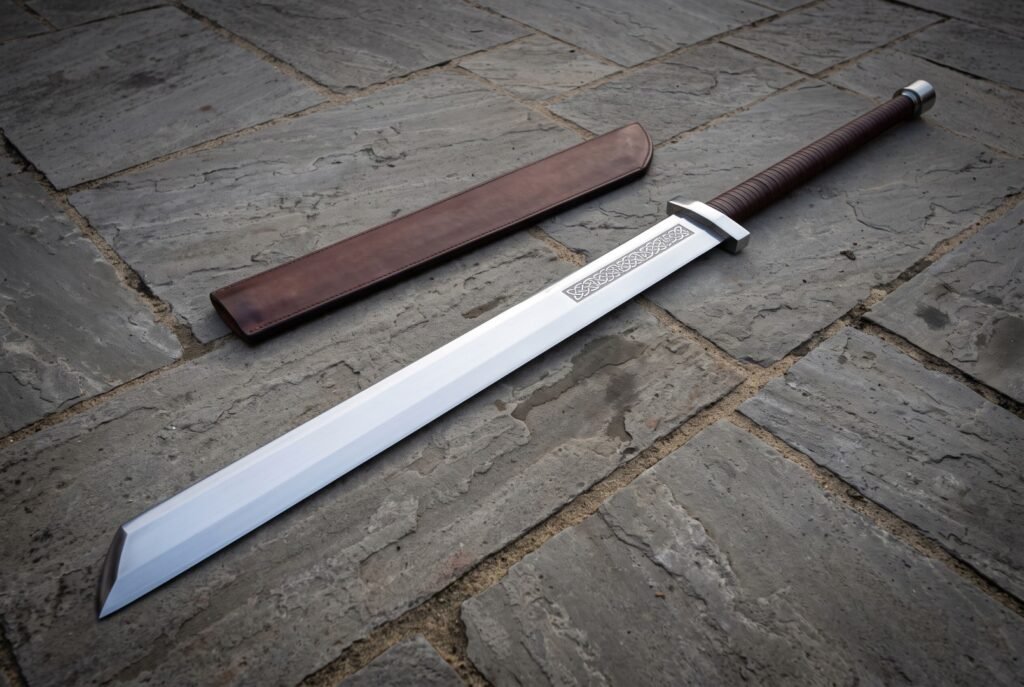 Broad single edged Viking langseax with a broken back blade, leather wrapped grip and brown scabbard lying on grey flagstones in soft daylight