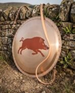 Bronze faced round hoplon shield with painted red boar leaning against a dry stone wall, tan leather carry strap draped over the face