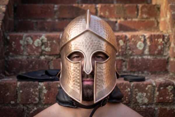 Bronze tone Corinthian Greek helmet with a hammered finish and Greek key border worn by a man against a brick wall