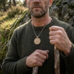 Bronze tone valknut pendant on a steel box chain worn over a green wool jumper by a man holding a wooden staff