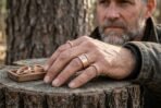 Bronze tone valknut stamped bands and twisted wire rings worn on a bearded man's hand resting on a tree stump with a walnut tray