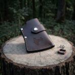 Brown leather axe head sheath with steel snaps and a tooled hammer motif standing on a tree stump, spare rivets beside it