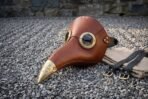 Brown leather plague doctor mask with brass goggle rims and engraved brass beak tip on grey gravel with straps