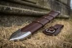 Brown leather sword scabbard with steel chape and brass rivets lying on dry grass beside its buckled belt frog, low angle