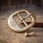 Brushed brass belt buckle with vegvisir engraving laid flat on stone beside a brown leather belt strap and two screw studs