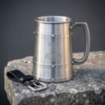 Brushed stainless steel viking tankard etched with a Mjolnir hammer and set with riveted bands, standing on a stone slab beside its black leather belt hanger