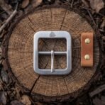 Brushed steel belt buckle with stamped Valknut and tan leather snap tab with brass studs, seen from above on a log