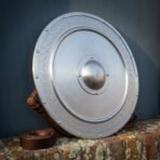 Brushed steel round shield tilted against a wall with its leather carry strap looped behind and coiled beside it