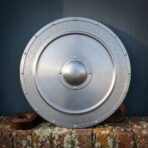 Brushed steel round shield with an engraved Celtic knotwork border, ringed rivets and a domed centre boss standing on a lichened brick wall beside a rolled leather strap