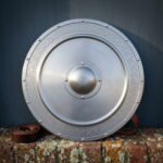 Brushed steel round shield with an engraved Celtic knotwork border, ringed rivets and a domed centre boss standing on a lichened brick wall beside a rolled leather strap