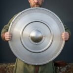 Brushed steel round shield with domed centre boss, stepped rings and knotwork band held up in both hands by a man in green