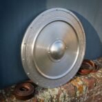 Brushed steel round shield with engraved knotwork border, riveted rim and domed boss leaning on a brick wall by a leather strap